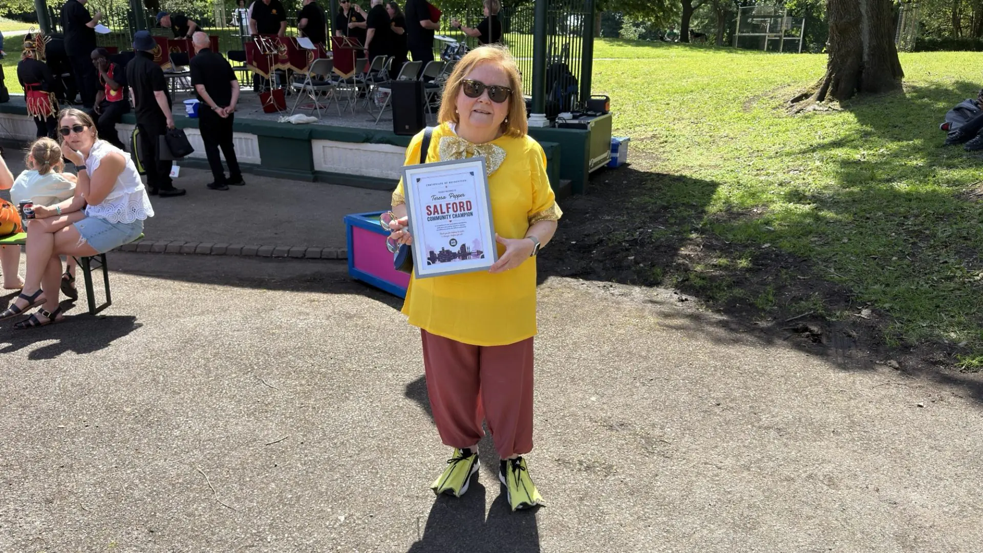 Woman in a bright yellow blouse and sunglasses holds a framed ‘Salford Community Champion’ certificate at a park event, with a band on a stage in the background.