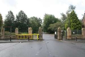 Entrance to Weaste Cemetery with a yellow barrier gate and stone pillars along a paved street.