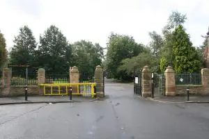 Entrance to Weaste Cemetery with a yellow barrier gate and stone pillars along a paved street.