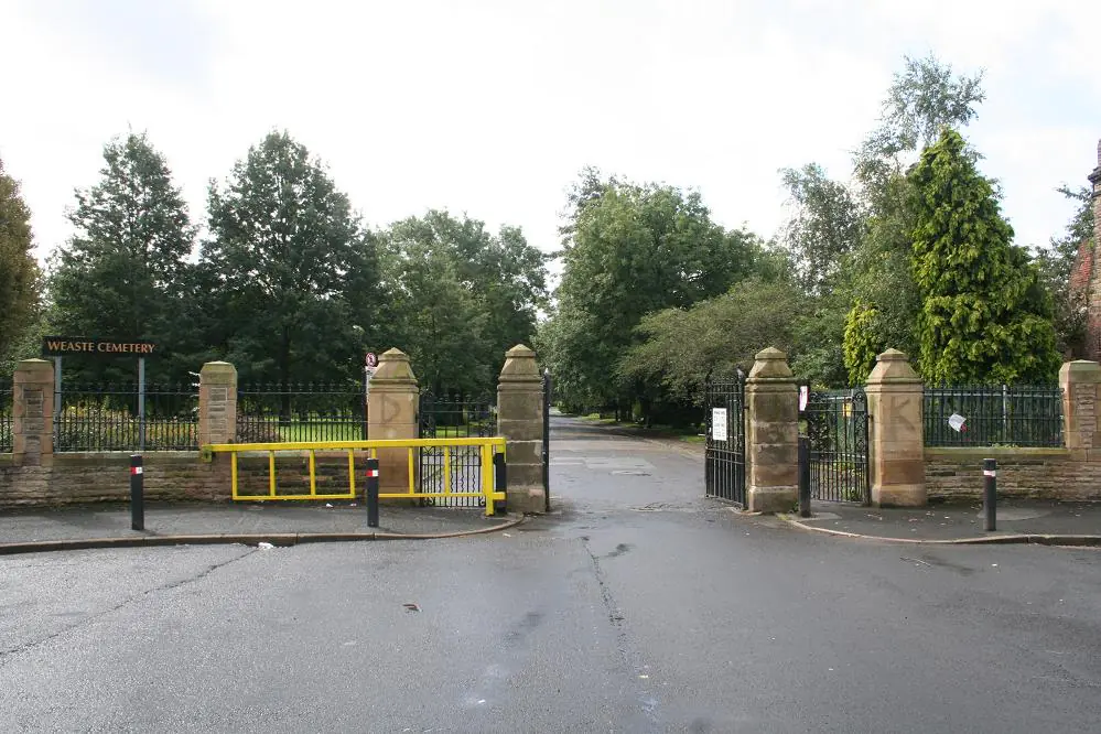 Entrance to Weaste Cemetery with a yellow barrier gate and stone pillars along a paved street.